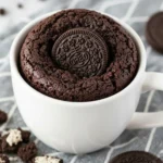 A rich chocolate Oreo mug cake served in a white ceramic mug, with crushed Oreo pieces visible.